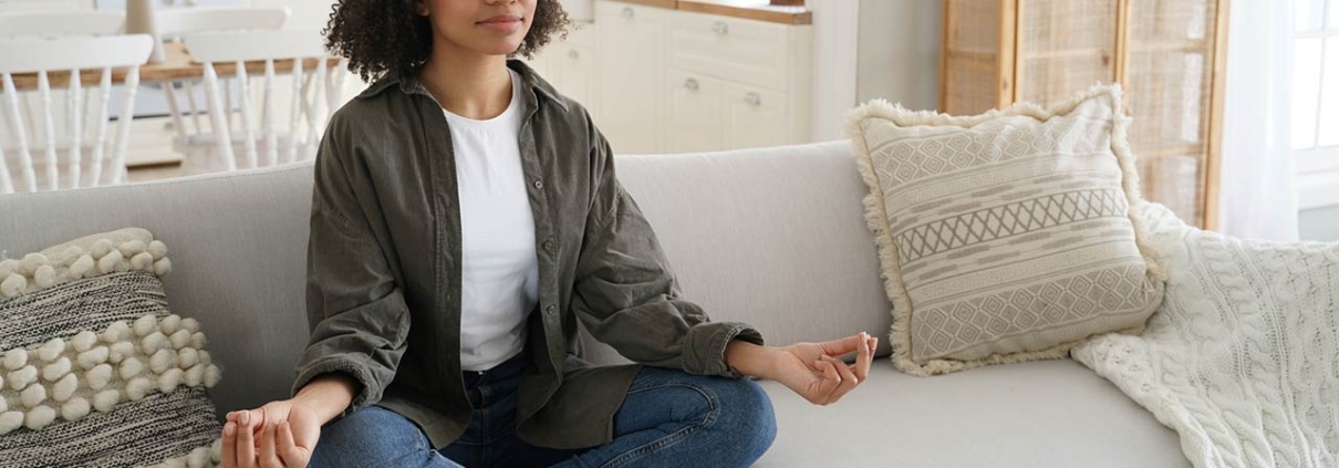 A serene Black woman meditates on a couch, eyes closed, in a sunlit, stylish living room